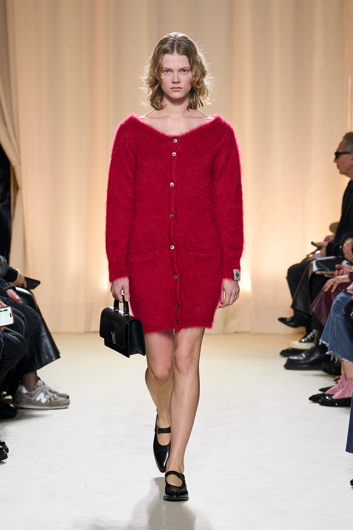 Model walks the runway in a bright red fuzzy cardigan buttoned down the front, carrying a small black handbag.