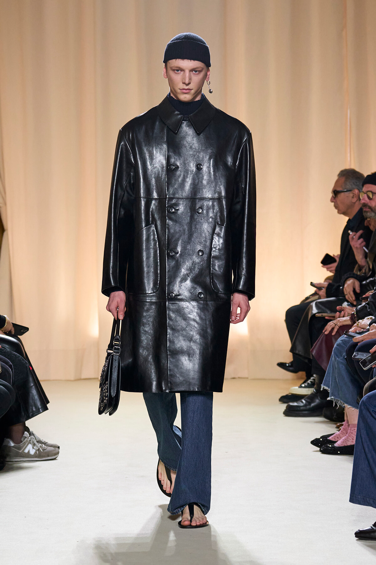 Model on a fashion runway wearing a glossy black double-breasted leather coat, black beanie, blue jeans, and flip-flop sandals, carrying a black handbag.