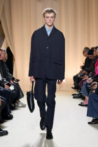 Male model walks the runway in a navy suit with a light blue shirt, holding a black handbag while audience watches from both sides.