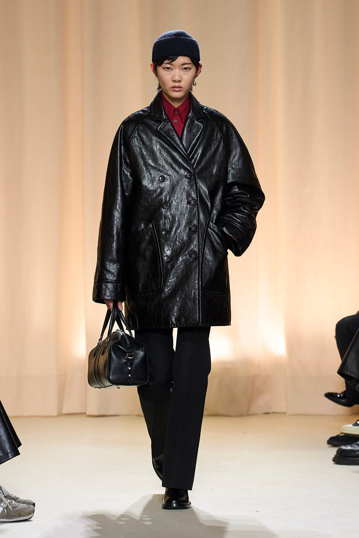 Model walking the runway in an oversized black leather double-breasted coat, red shirt, black pants, beanie, and a black handbag.