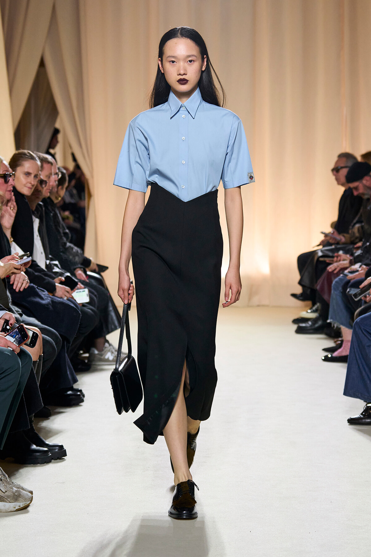 Model walks a runway in a light blue short-sleeve button-up shirt with a black high-waisted skirt, carrying a black bag. Audience seated on both sides.
