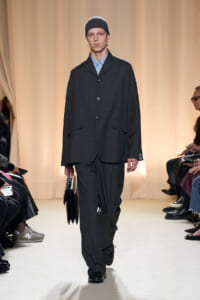 Male model on a runway wearing a dark oversized suit and a black knit beanie, carrying a black bag.