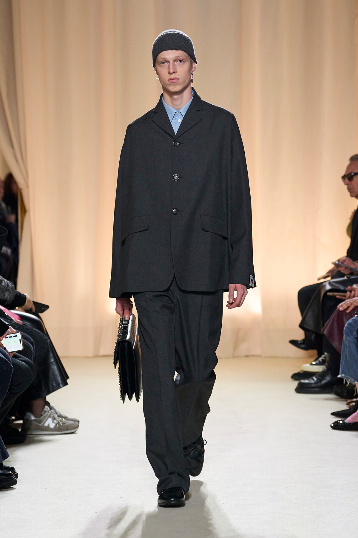 Male model on a runway wearing a dark oversized suit and a black knit beanie, carrying a black bag.
