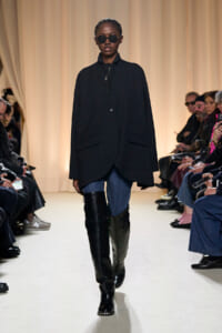 Model walks runway in oversized black coat, round sunglasses, and tall black boots; cream backdrop and seated audience visible on sides
