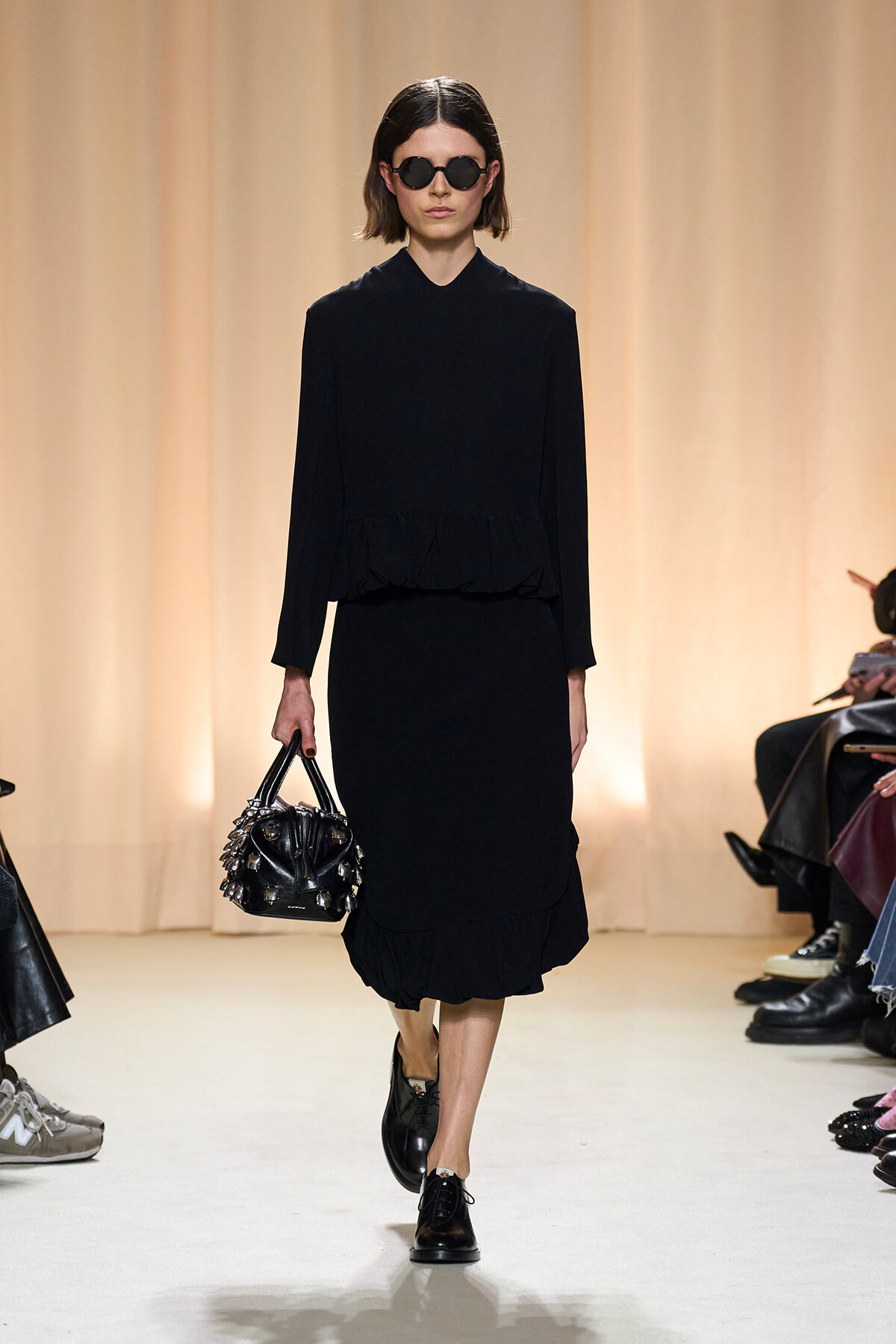 Model walks the runway in a black, long-sleeve dress with a ruffled hem, wearing round sunglasses and carrying a spiked black handbag.