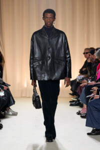 Male model on a runway wearing a glossy black leather blazer with black trousers, carrying a small studded black handbag; beige curtain backdrop.