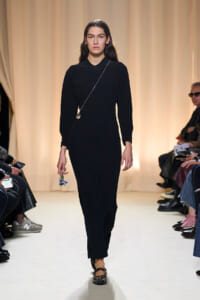Model walks the runway in a black long-sleeve sheath dress, holding a small bouquet in her left hand and a thin crossbody chain necklace visible.