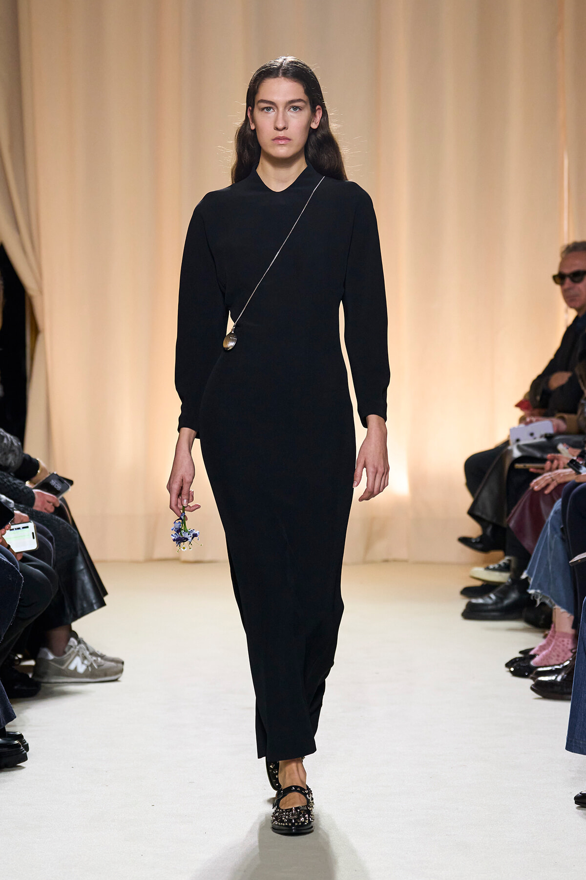 Model walks the runway in a black long-sleeve sheath dress, holding a small bouquet in her left hand and a thin crossbody chain necklace visible.