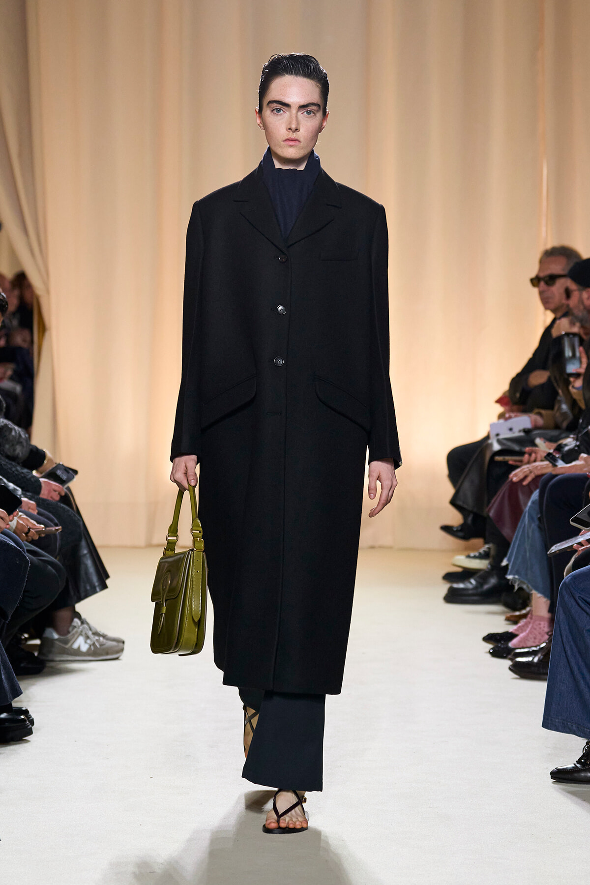 Model walks the runway in a long black coat over a dark blue high-neck top, carrying an olive green handbag, audience visible on sides.