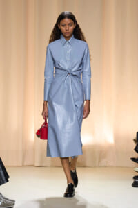 Model walks runway in a shiny light-blue midi dress with a front waist knot, pairing it with a red handbag.