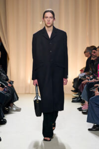 Model walks a runway in a long black coat, carrying a black handbag, with a beige curtain backdrop and seated spectators on the sides.