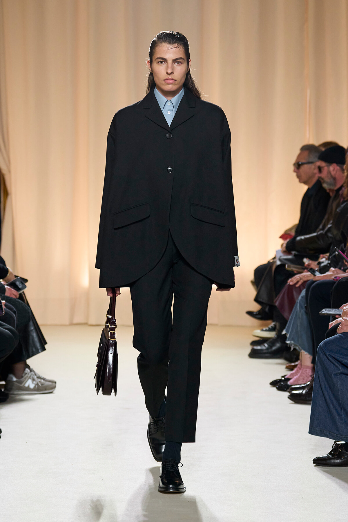 Model walking the runway in an oversized black suit with a light blue shirt, carrying a dark brown handbag as audience watches from the sides.