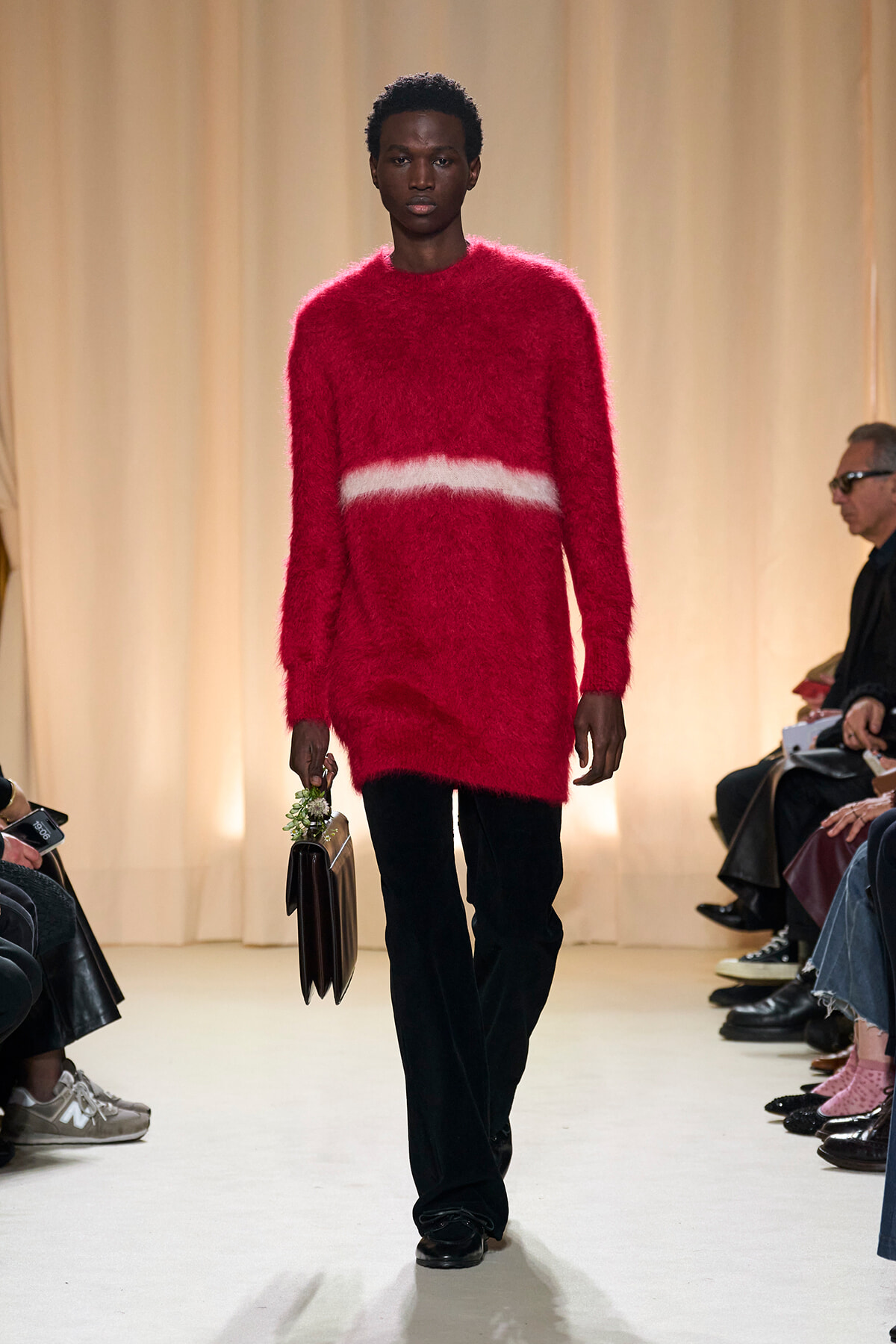 Male model on a runway wearing a bright red fuzzy sweater with a white stripe and black pants, carrying a dark handbag.