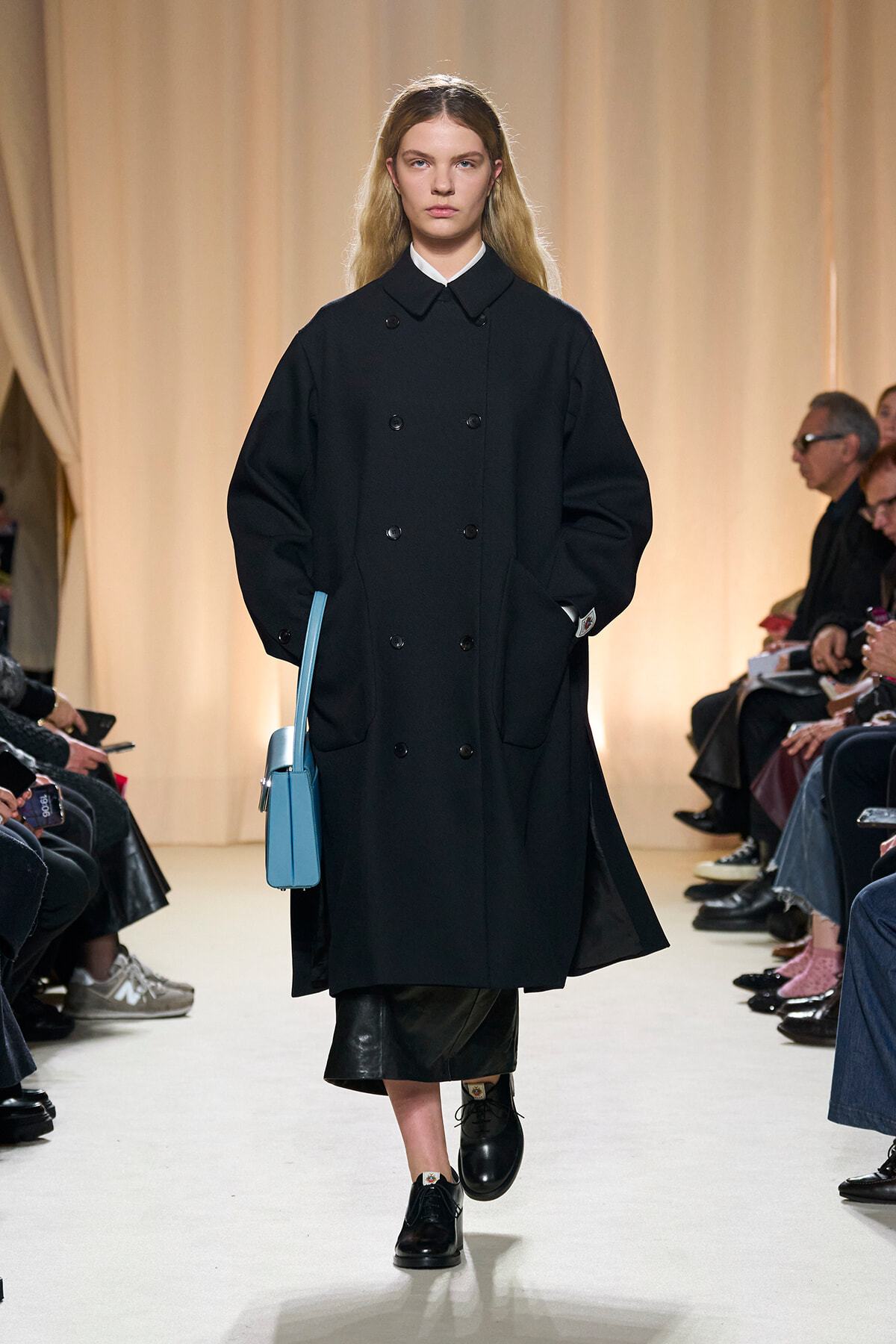 Model walking the runway in a long black double-breasted coat with a light-blue handbag at her side.