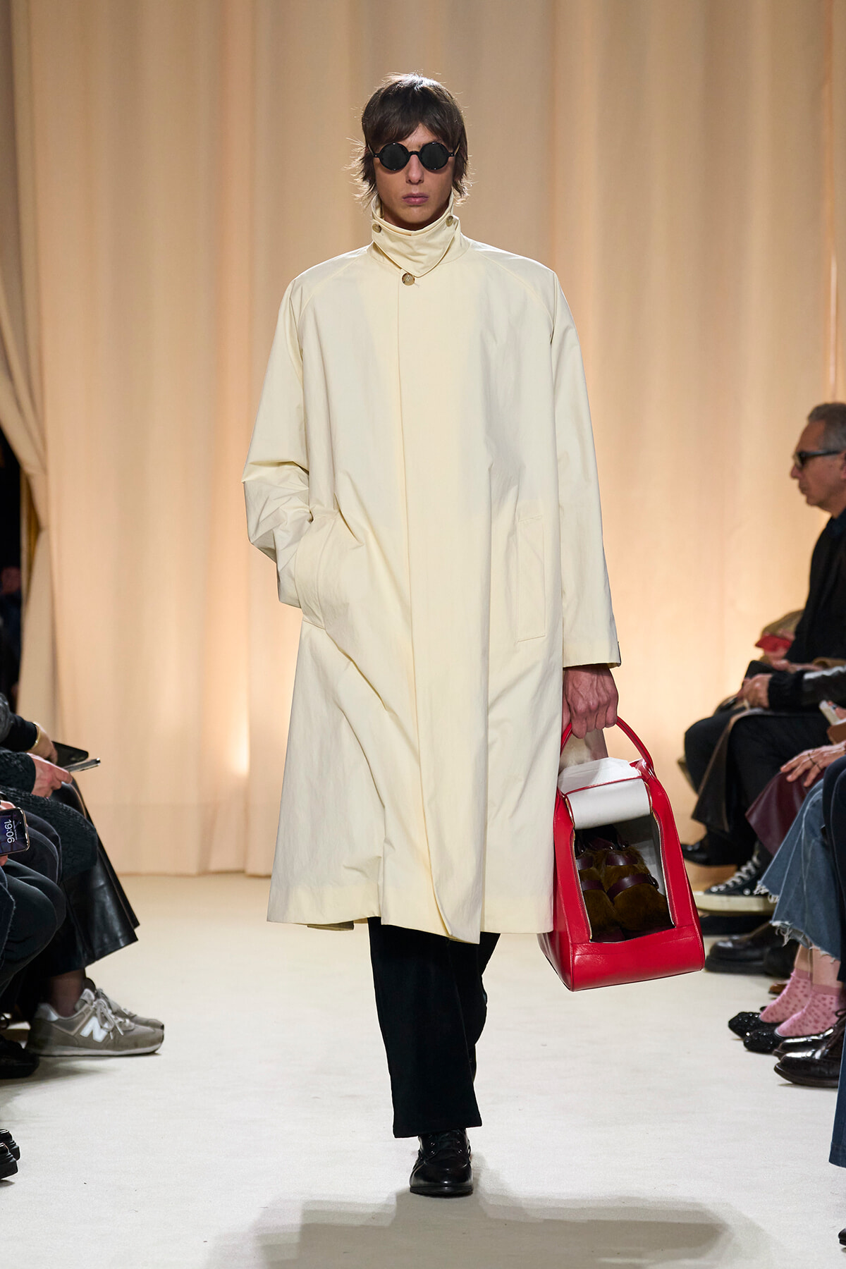 Model on a runway wearing a cream oversized coat, round black sunglasses, and carrying a red-and-white handbag by her side.