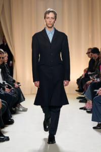 Model walking a fashion show runway in a knee-length black coat over a light blue button-down shirt; audience seated along the sides of the catwalk.