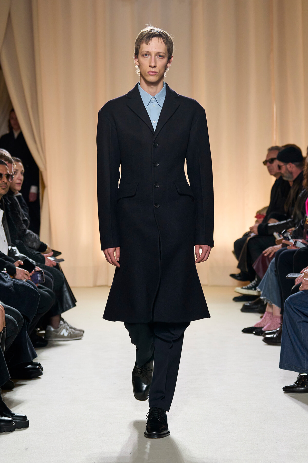 Model walking a fashion show runway in a knee-length black coat over a light blue button-down shirt; audience seated along the sides of the catwalk.