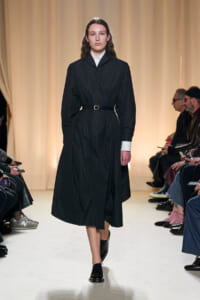 Model walks the runway in a black belted trench dress with white cuffs and collar, audience seated on sides.