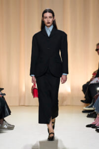 Model walking a runway in a black tailored suit with a light blue shirt and a red handbag tucked under her arm while spectators watch from the sides.