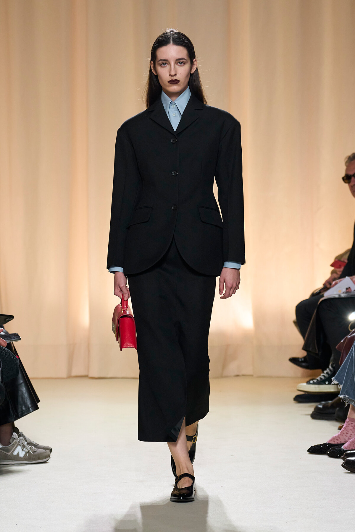 Model walking a runway in a black tailored suit with a light blue shirt and a red handbag tucked under her arm while spectators watch from the sides.
