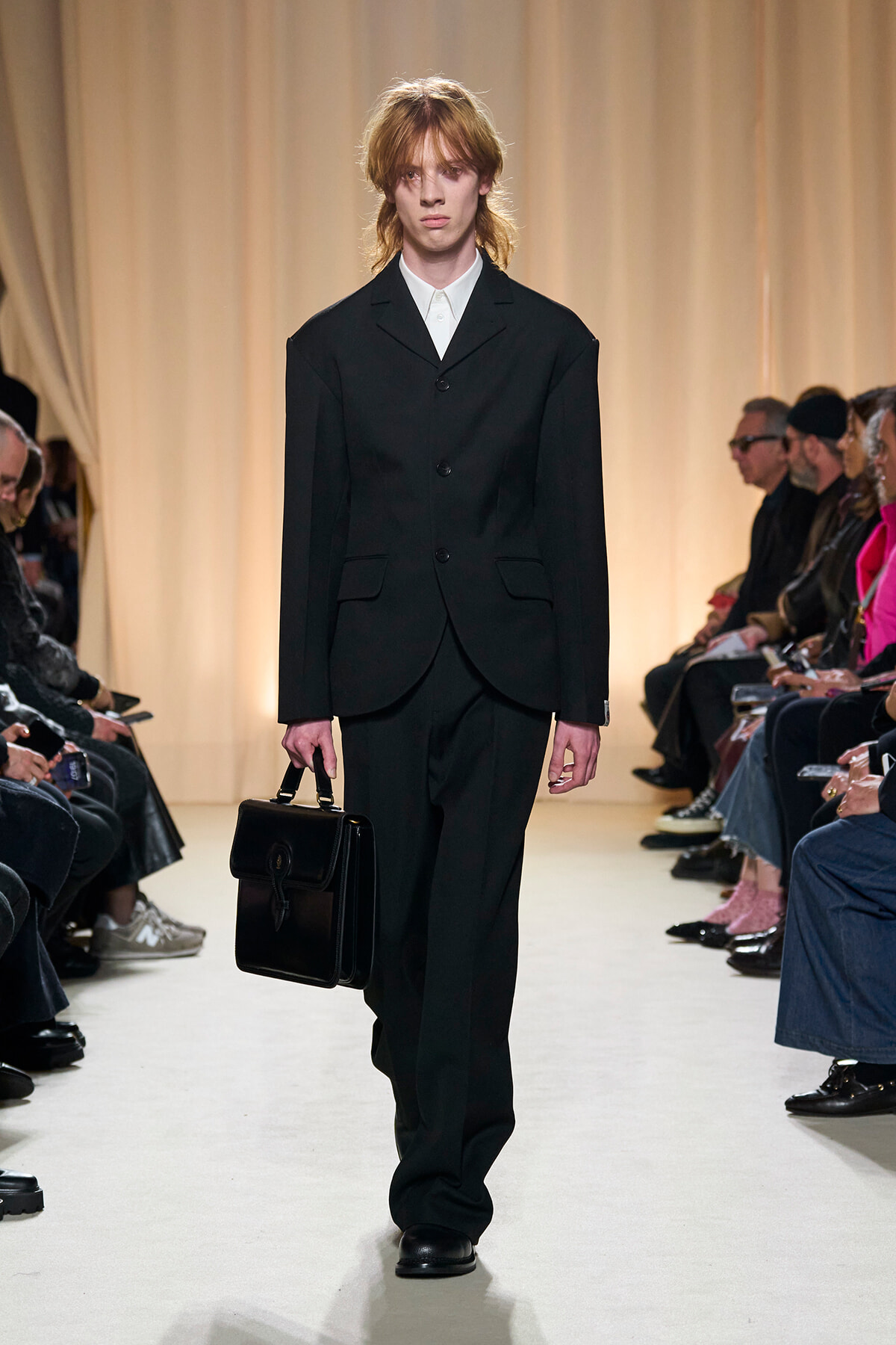 Male model walks a fashion show in a black tailored suit with a white shirt, carrying a black briefcase on a beige-lit runway