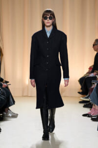 Model on a runway wearing a long black buttoned coat, light blue shirt collar, round sunglasses, and knee-high lace-up boots.
