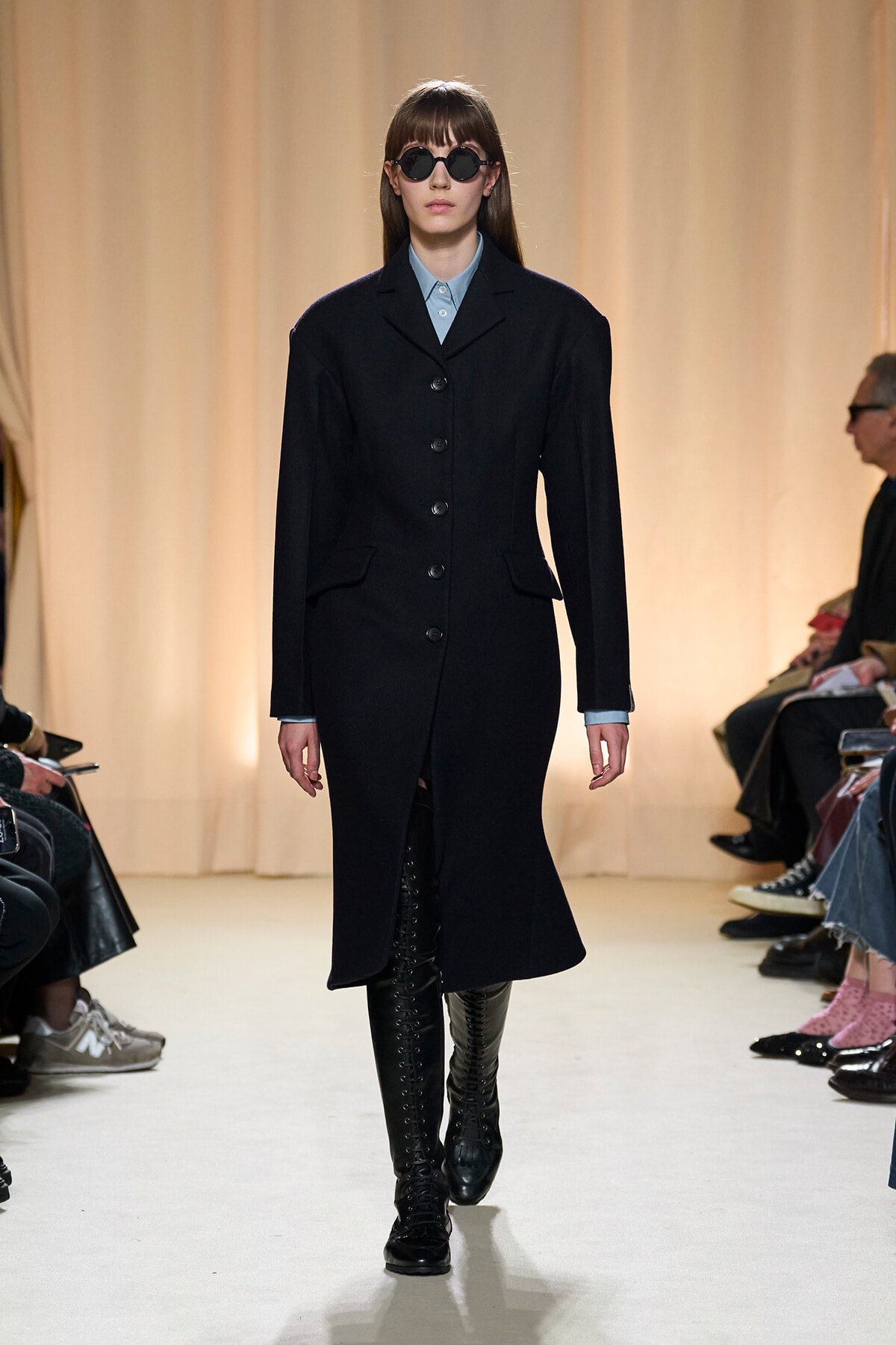 Model on a runway wearing a long black buttoned coat, light blue shirt collar, round sunglasses, and knee-high lace-up boots.