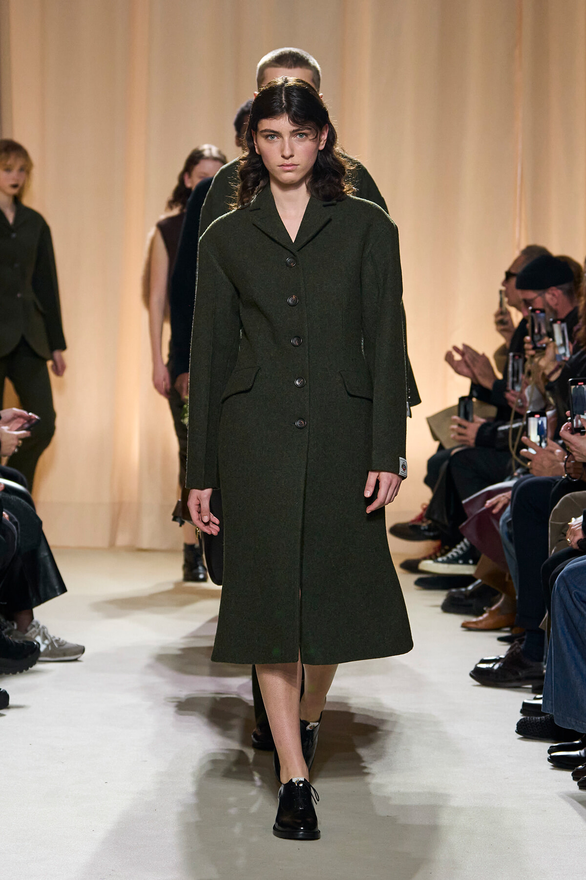 Model walks a fashion runway wearing a dark green knee-length wool coat with large buttons, black shoes, and a focused expression; audience and photographers line the sides under warm lighting.