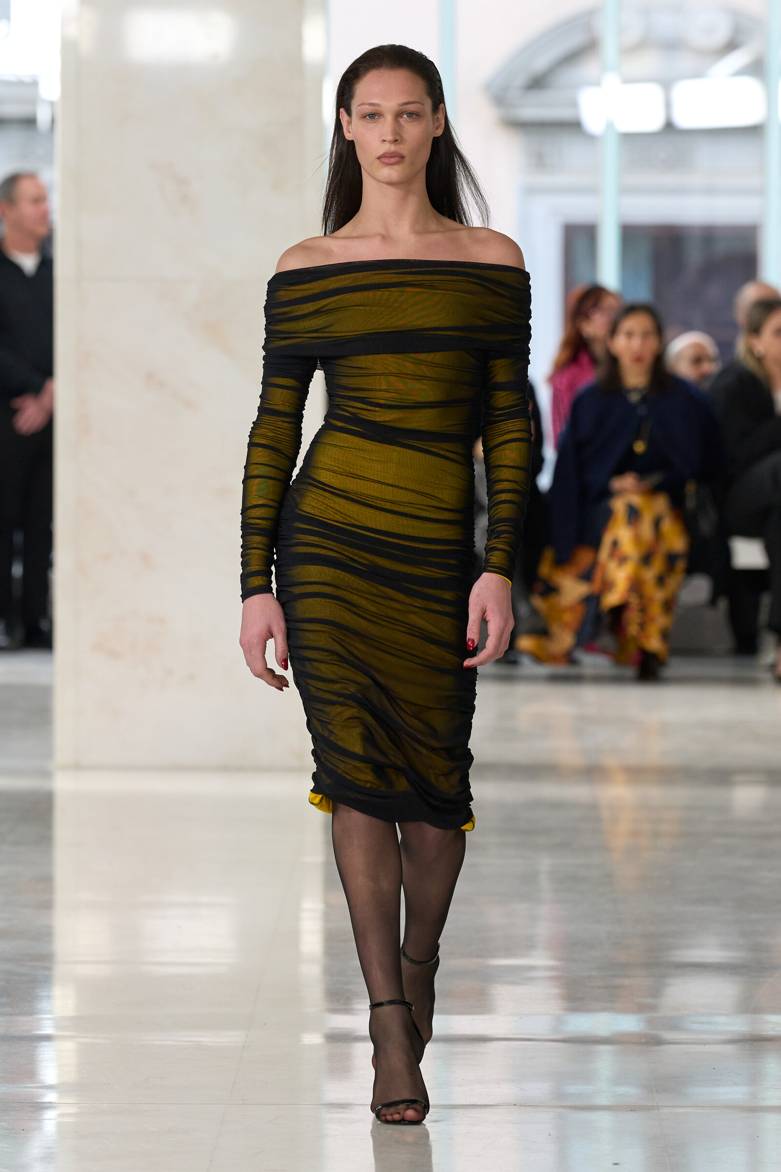 Model walks down a runway in a gold and black off-the-shoulder striped dress, audience blurred in the background.