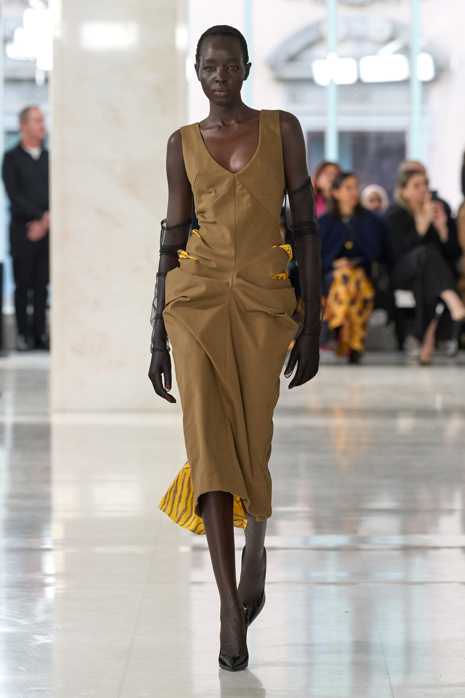 Model walks a fashion runway wearing a tan, structured dress with yellow accents and sheer black sleeves; blurred audience in the background.