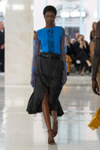 Model walks a fashion runway wearing a bright blue sleeveless top with sheer blue sleeves and a black belted skirt; blurred audience in background.