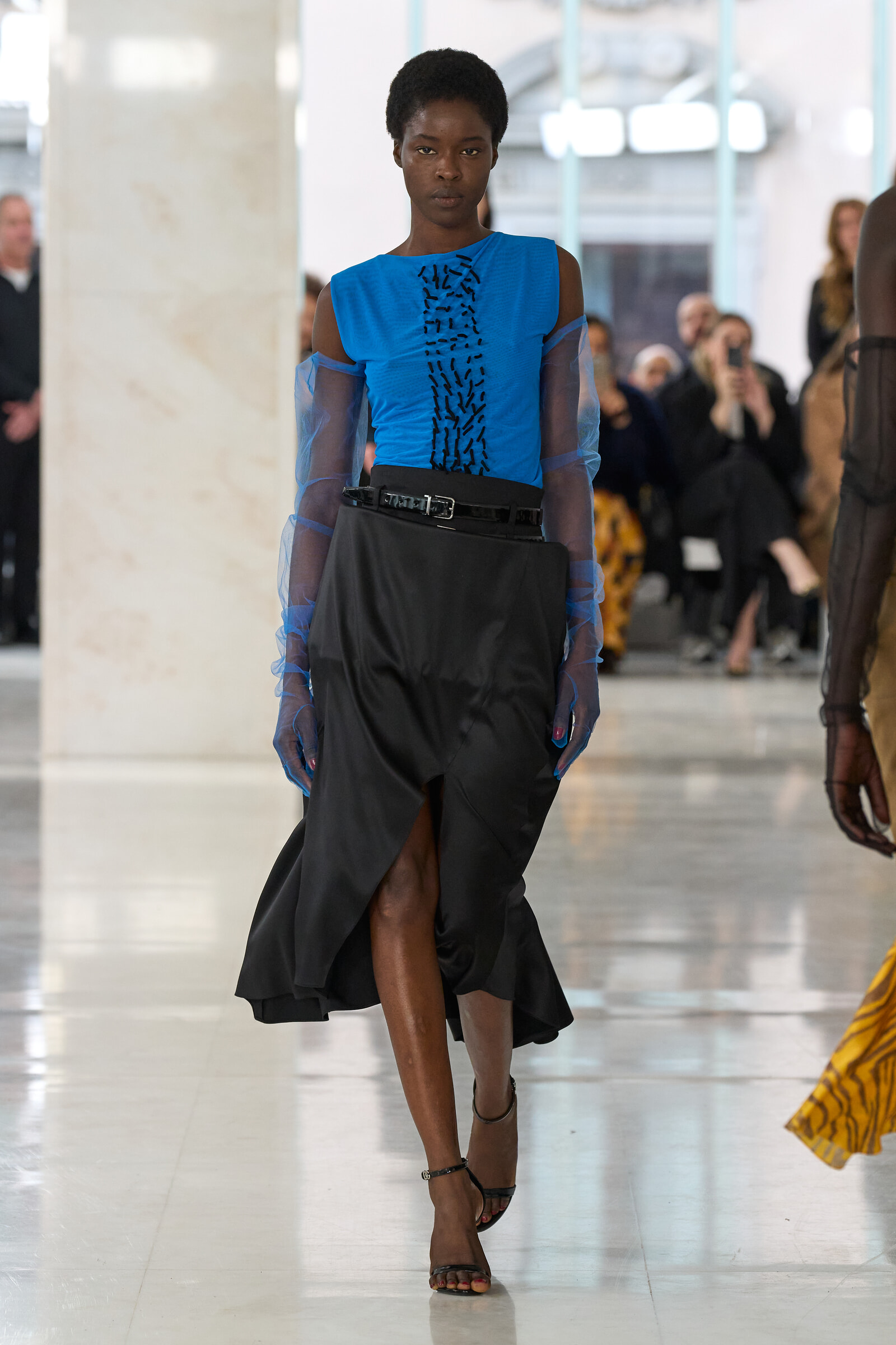 Model walks a fashion runway wearing a bright blue sleeveless top with sheer blue sleeves and a black belted skirt; blurred audience in background.