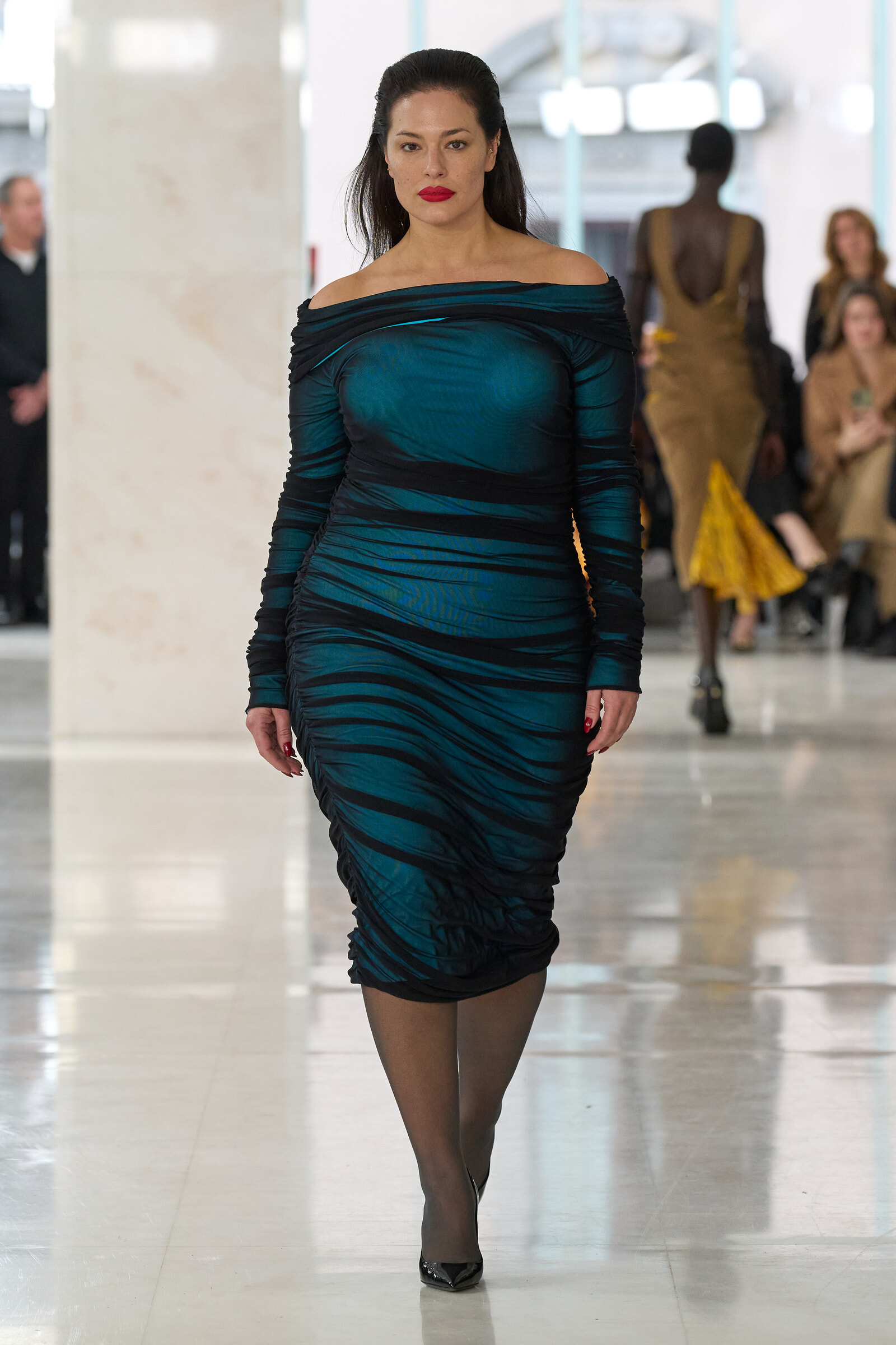 Model walking confidently on a fashion runway in a teal off-shoulder, ruched dress with black stripes and red lipstick.