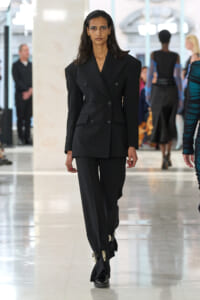 Model walking a fashion runway in a black double-breasted suit with wide shoulders, focused expression.