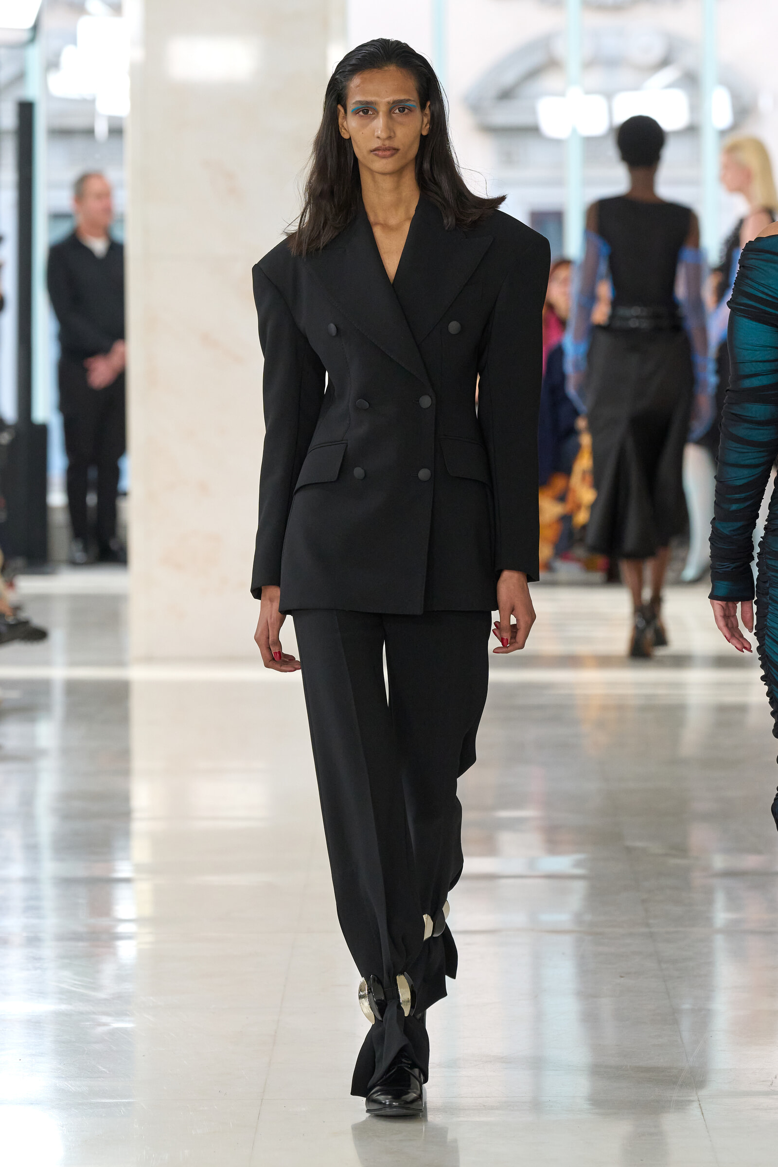 Model walking a fashion runway in a black double-breasted suit with wide shoulders, focused expression.