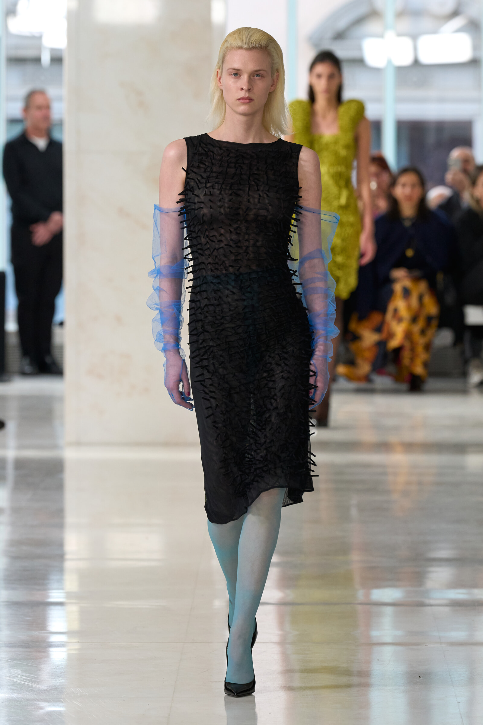 Model walking a fashion runway in a textured black dress with sheer blue sleeves; audience blurred in background.