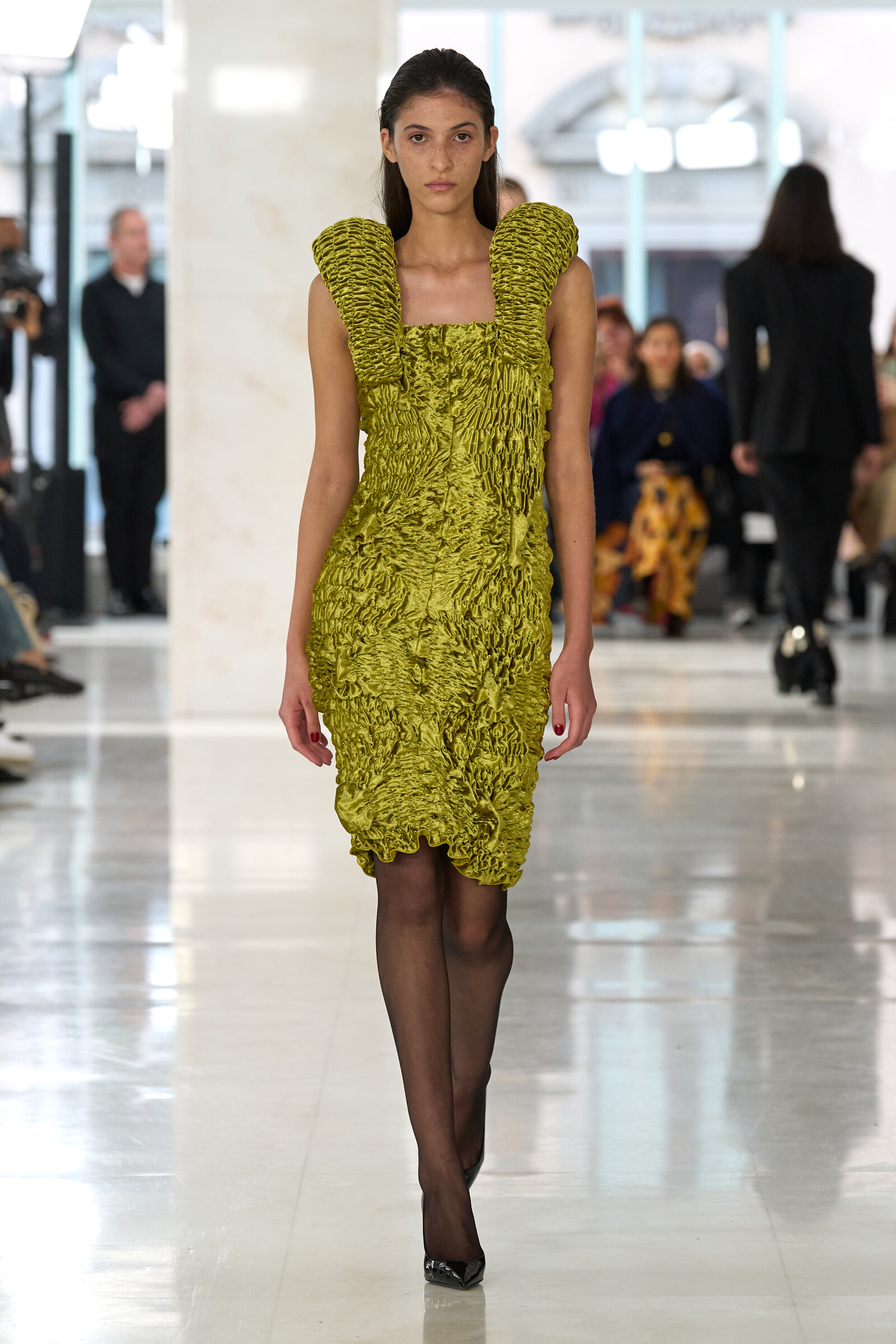 Model walking a fashion runway in a lime-green textured dress with sculpted shoulder details; audience blurred in background.