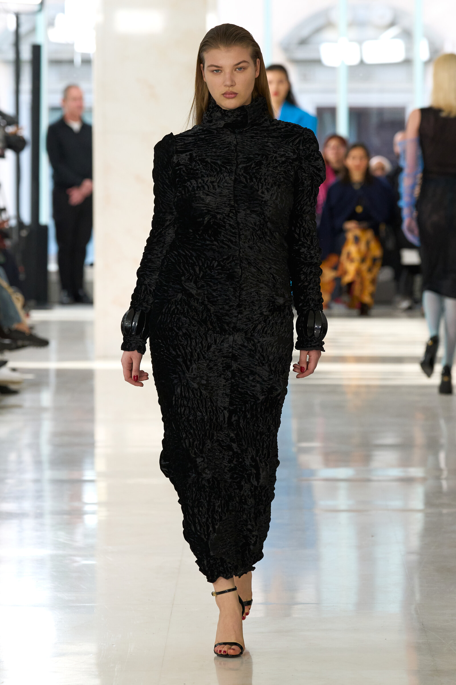 Model walking down a fashion runway in a black textured high-collar dress, audience blurred in the background.