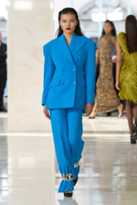 Model walking a fashion show wearing a bright blue, double-breasted suit with wide shoulders and matching trousers.