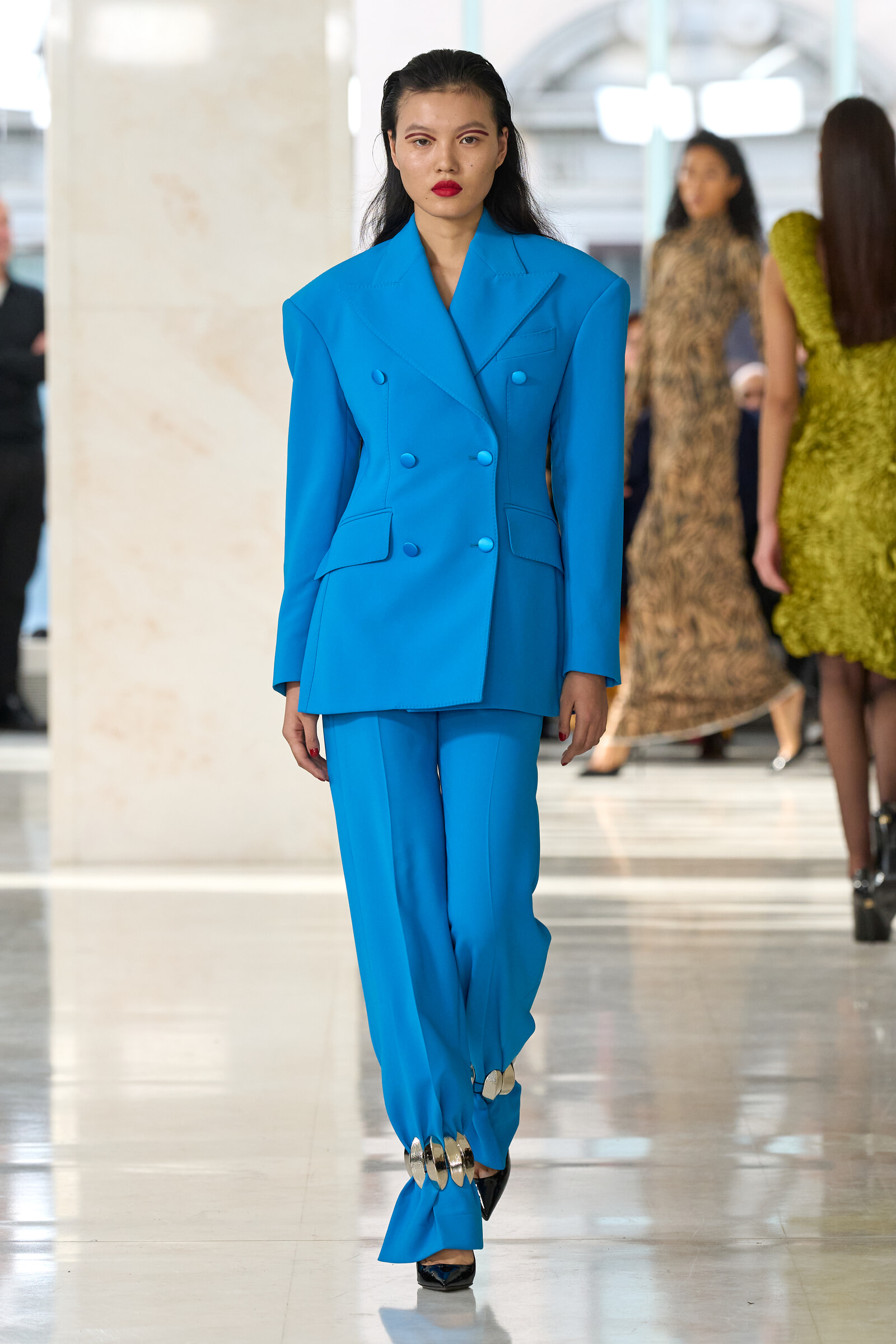 Model walking a fashion show wearing a bright blue, double-breasted suit with wide shoulders and matching trousers.