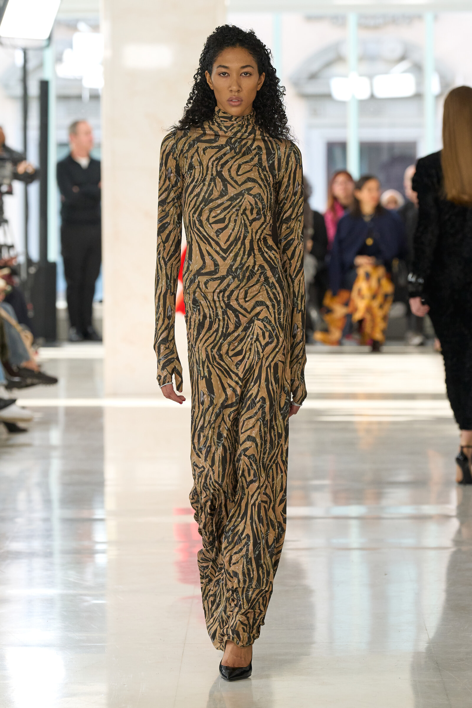 Female model walking a fashion runway in a brown and black tiger-print long-sleeve dress; blurred audience in background due to depth of field