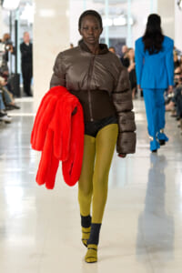 Model on a runway wearing a brown puffer jacket, mustard tights, and yellow sandals, holding a bright red fur coat.