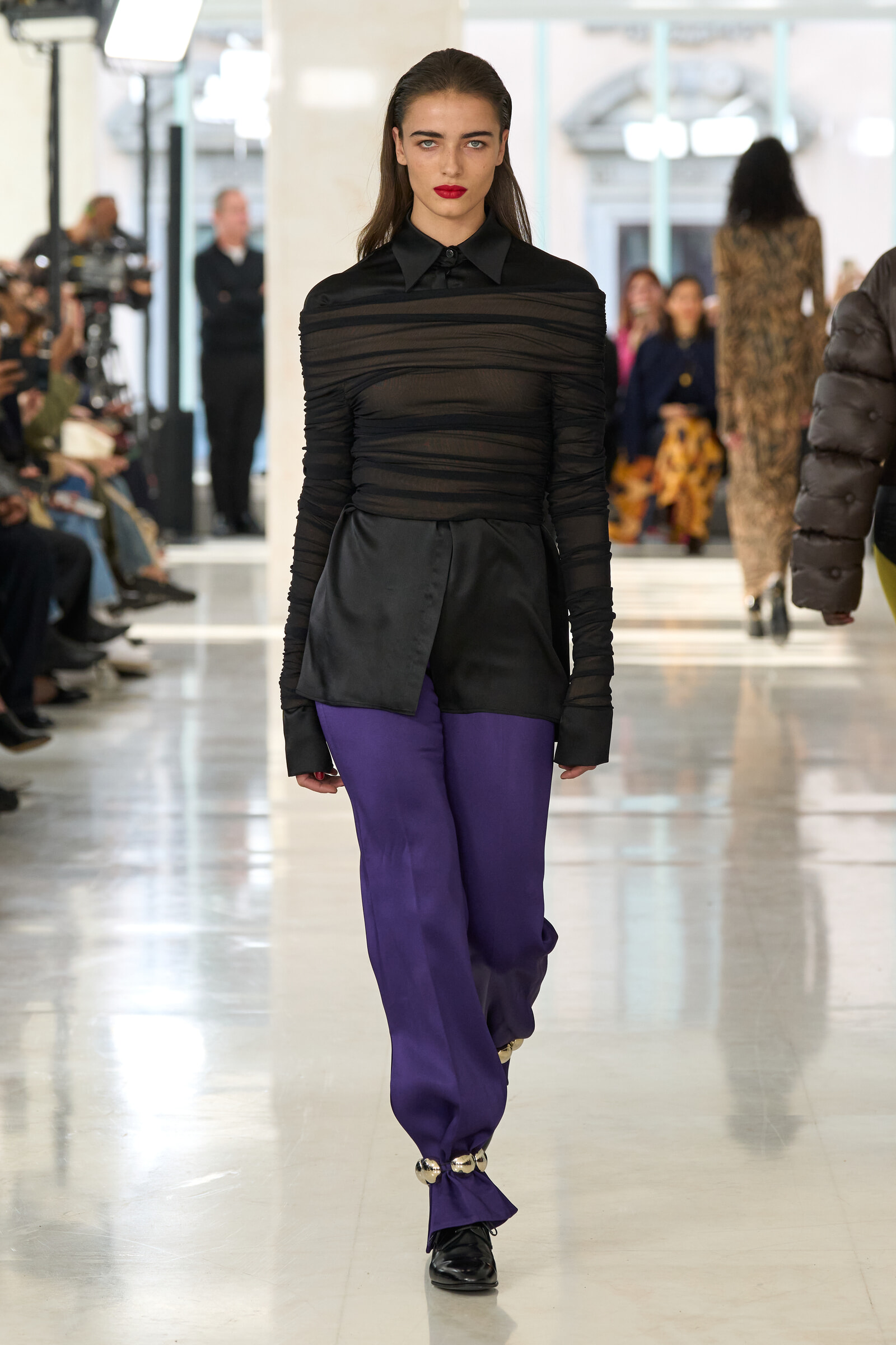 Model walking the runway in a sheer black top and purple pants, bold red lipstick, with photographers and audience visible in a bright venue.