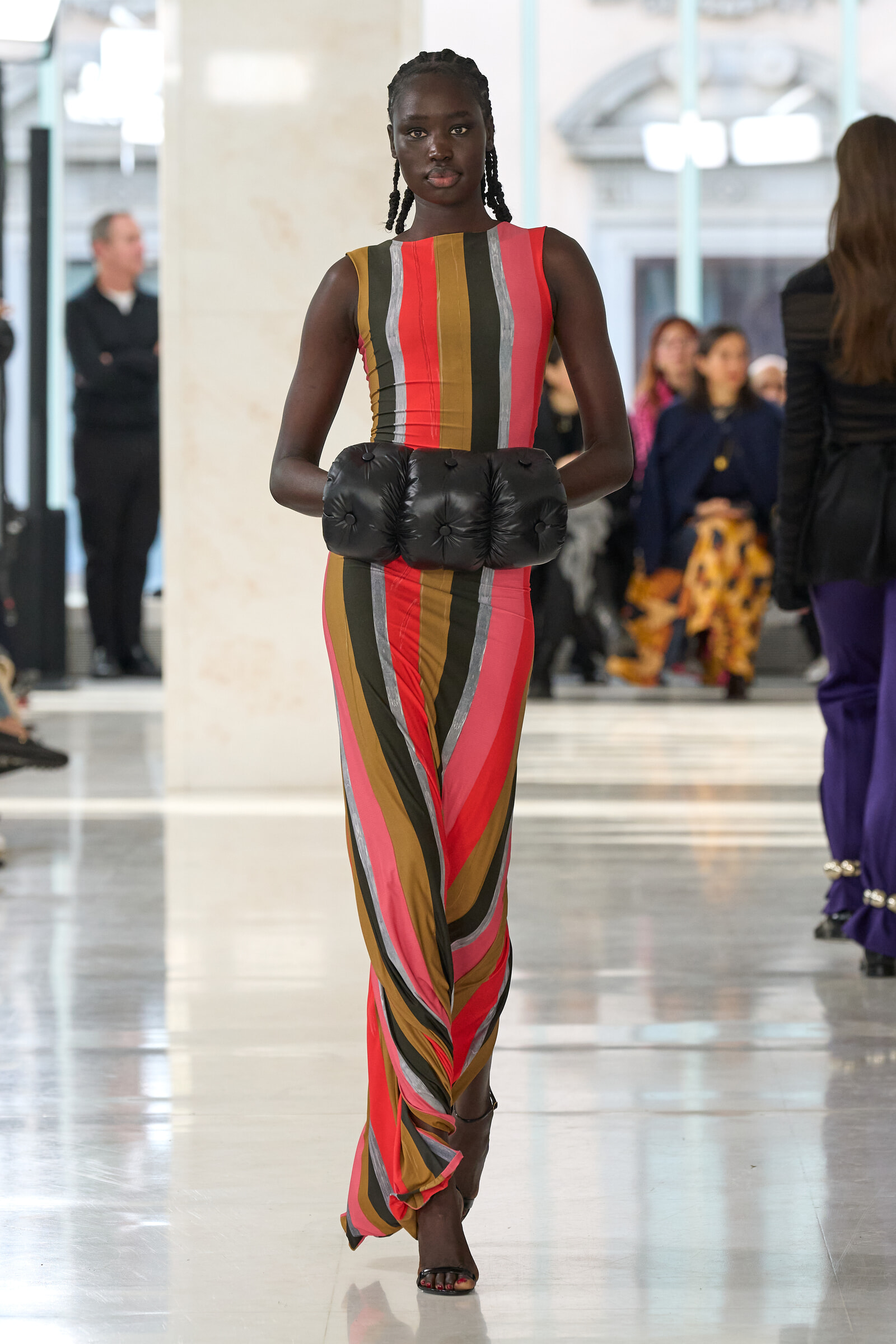 Model walking the runway in a sleeveless, multicolored striped dress with oversized black padded gloves; fashion show setting in background.