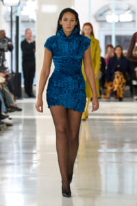 Model walking a blue ruching mini dress on a fashion-show runway, focused expression, blurred audience in background