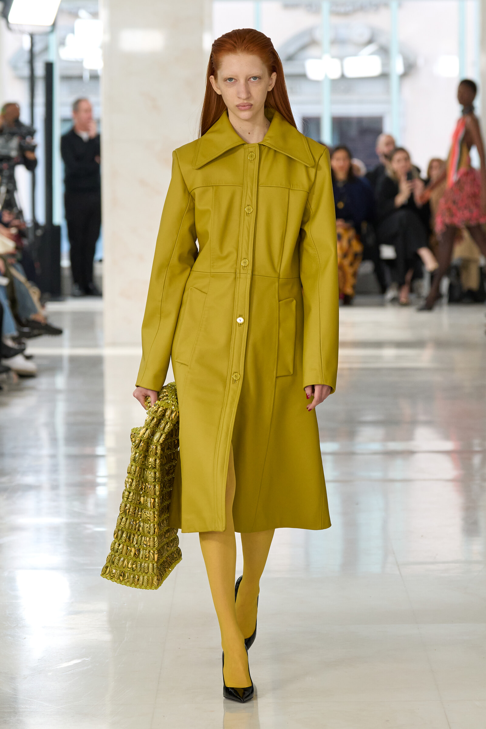 Model walking a fashion runway in a mustard-yellow coat, matching tights, black heels, and a textured gold bag in hand.