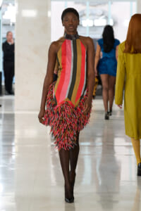 Model walking the runway in a sleeveless, multicolored striped dress with fringe at the hem.