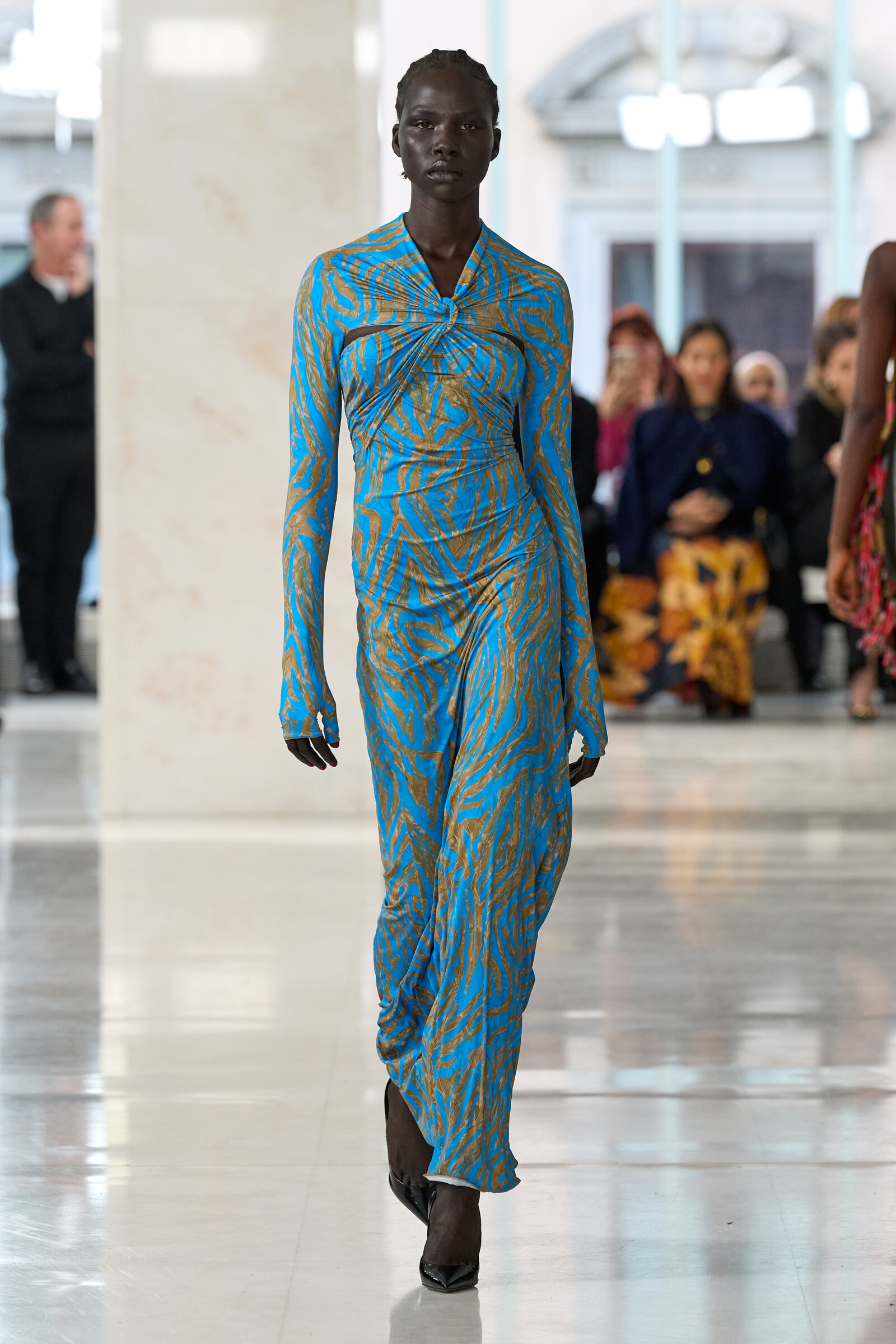 Full-body view of a fashion model walking the runway in a blue and tan zebra-print dress, with blurred audience in the background.
