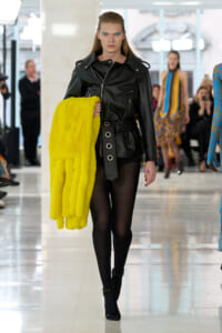 Model on a fashion runway wearing a black leather jacket and sheer tights, holding a bright yellow fur coat.