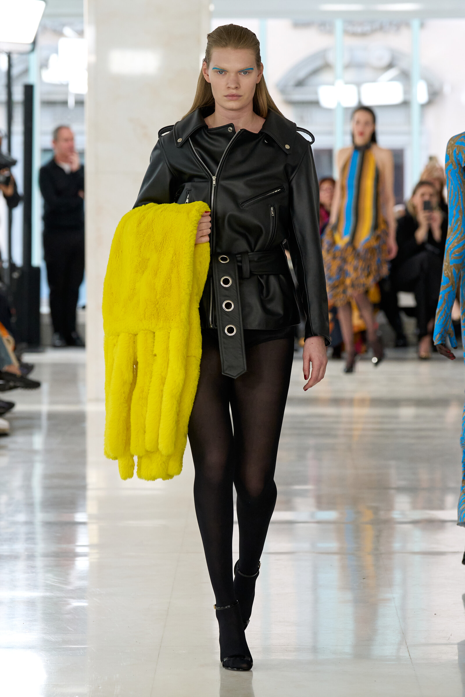 Model on a fashion runway wearing a black leather jacket and sheer tights, holding a bright yellow fur coat.